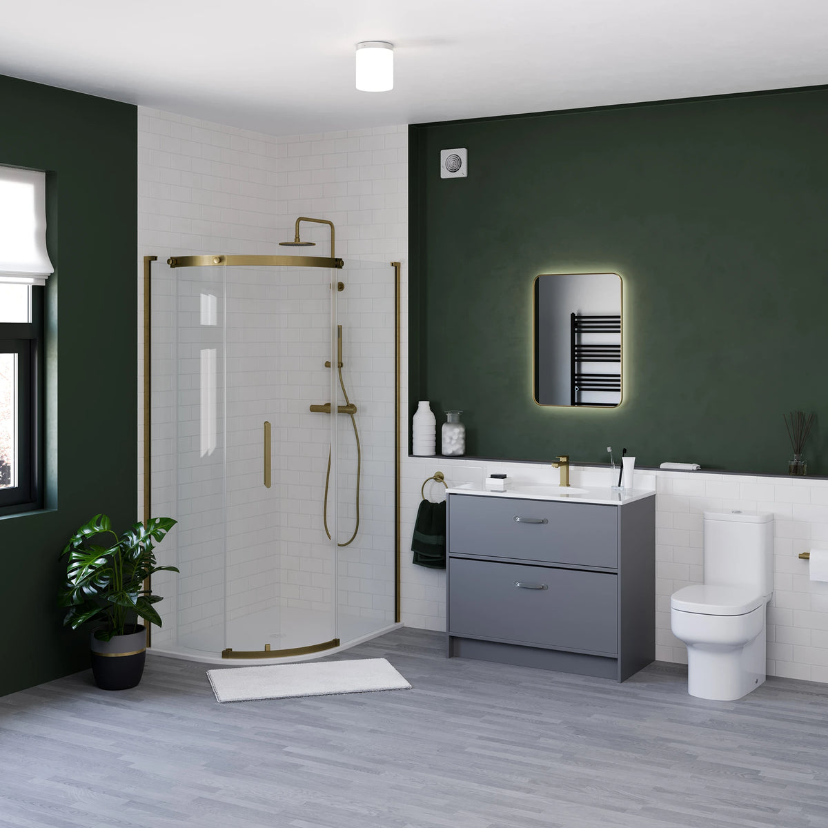 A modern bathroom interior featuring a glass shower screen with gold-toned hardware, a gray double-sink vanity, a white toilet, and a dark green accent wall with an illuminated mirror. #Finish_Brushed Brass
