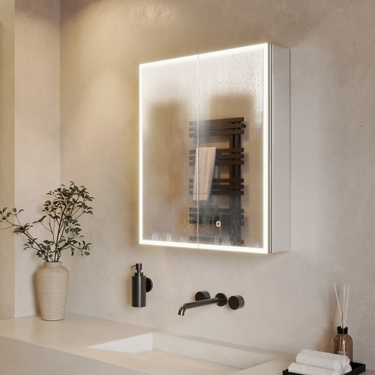A modern wall-mounted medicine cabinet with an illuminated LED light frame and frosted glass doors, installed above a bathroom sink with matte black fixtures. The cabinet reflects a dark wood heated towel rail. #Size_600mm x 700mm