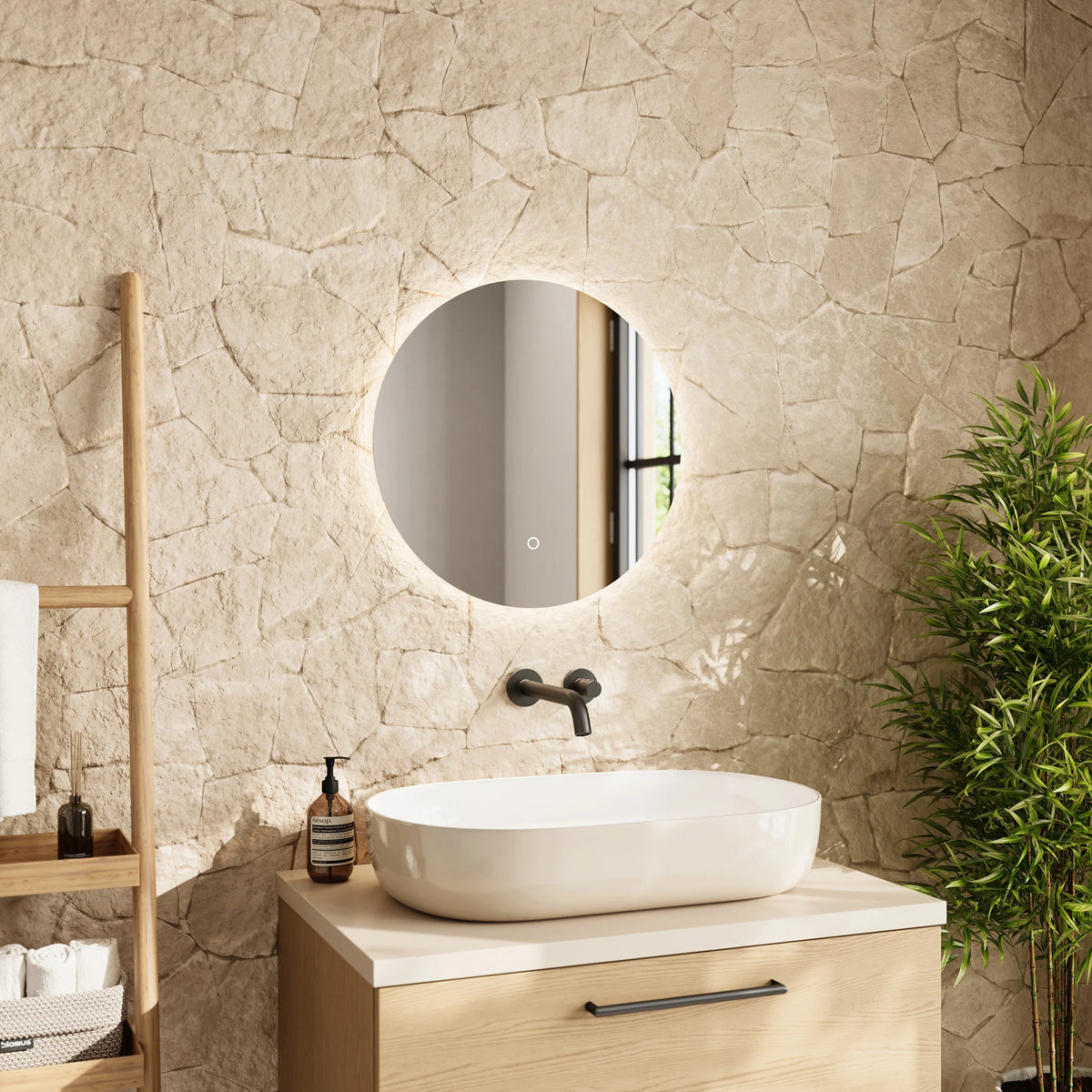 A modern bathroom vanity setup featuring a round backlit LED wall mirror mounted on a textured beige stone wall. Below the mirror is a white vessel sink on a light wood vanity cabinet. #Size_⌀500mm