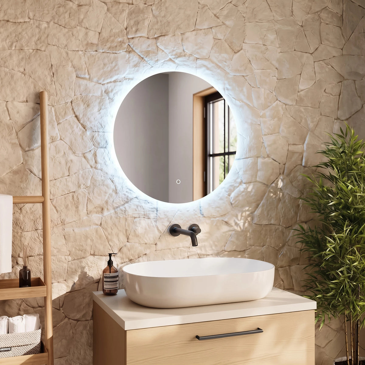 An illuminated round wall mirror with a modern matte black faucet and a white vessel sink on a light wood vanity, set against a textured stone wall with a wooden shelf and green plant. #Size_⌀600mm