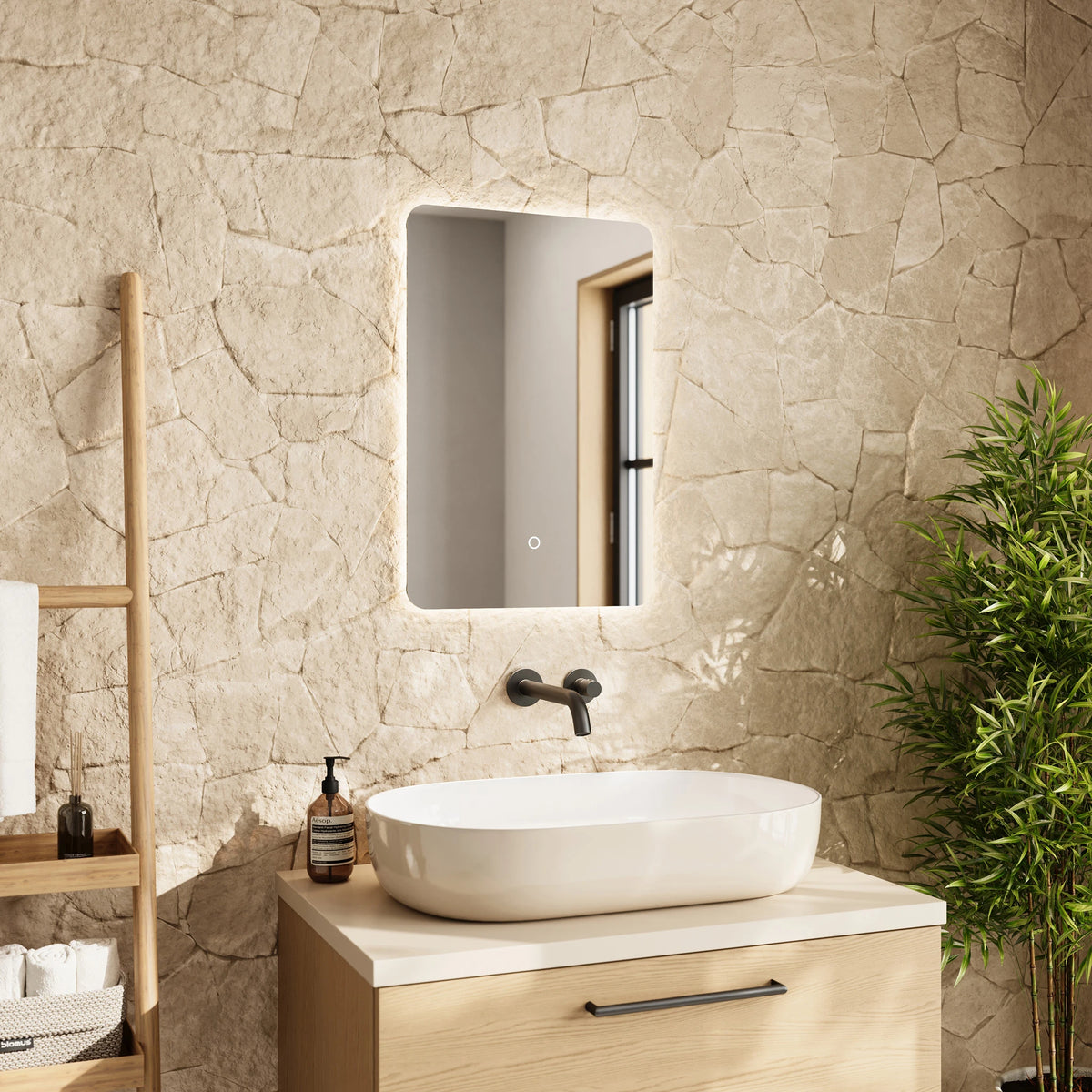 A modern bathroom scene featuring a rectangular LED-backlit mirror mounted on a textured beige stone wall above a light wood vanity with a white vessel sink. A wooden shelf with toiletries and a green potted plant are visible in the background. #Size_400mm x 600mm