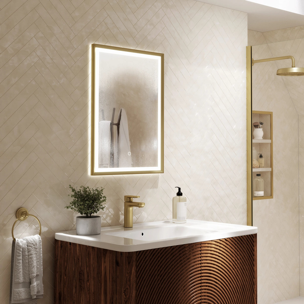 A modern bathroom vanity featuring a rectangular backlit LED mirror with a gold frame, a white countertop, a gold faucet, and a dark wood cabinet with a wavy front panel. The background consists of beige diamond-patterned tile walls, and a matching gold shower fixture is visible in the shower enclosure on the right. #Finish_Brushed Brass