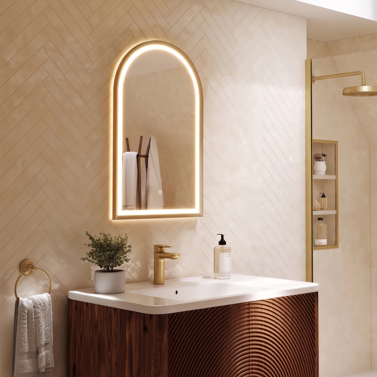 An illuminated arched wall mirror with a gold frame hangs above a white sink and a wood vanity in a modern bathroom. The wall has beige herringbone tile, and a gold shower fixture is visible to the right. #Finish_Brushed Brass