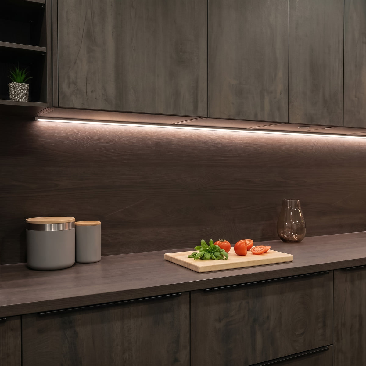 A modern kitchen scene with dark wood cabinets, a matching countertop, under-cabinet LED lighting, and a wooden cutting board with tomatoes and basil on the counter. #Finish_Black