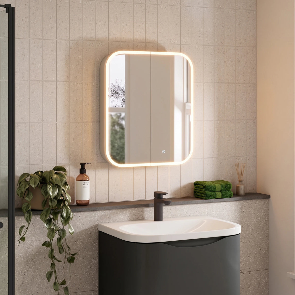 A modern bathroom scene featuring a rectangular wall-mounted mirror with a glowing warm white LED light border, installed above a white and dark gray pedestal sink. The background consists of light beige textured wall tiles, with a dark shelf holding a green towel, a plant, and a bottle. #Size_600mm x 700mm