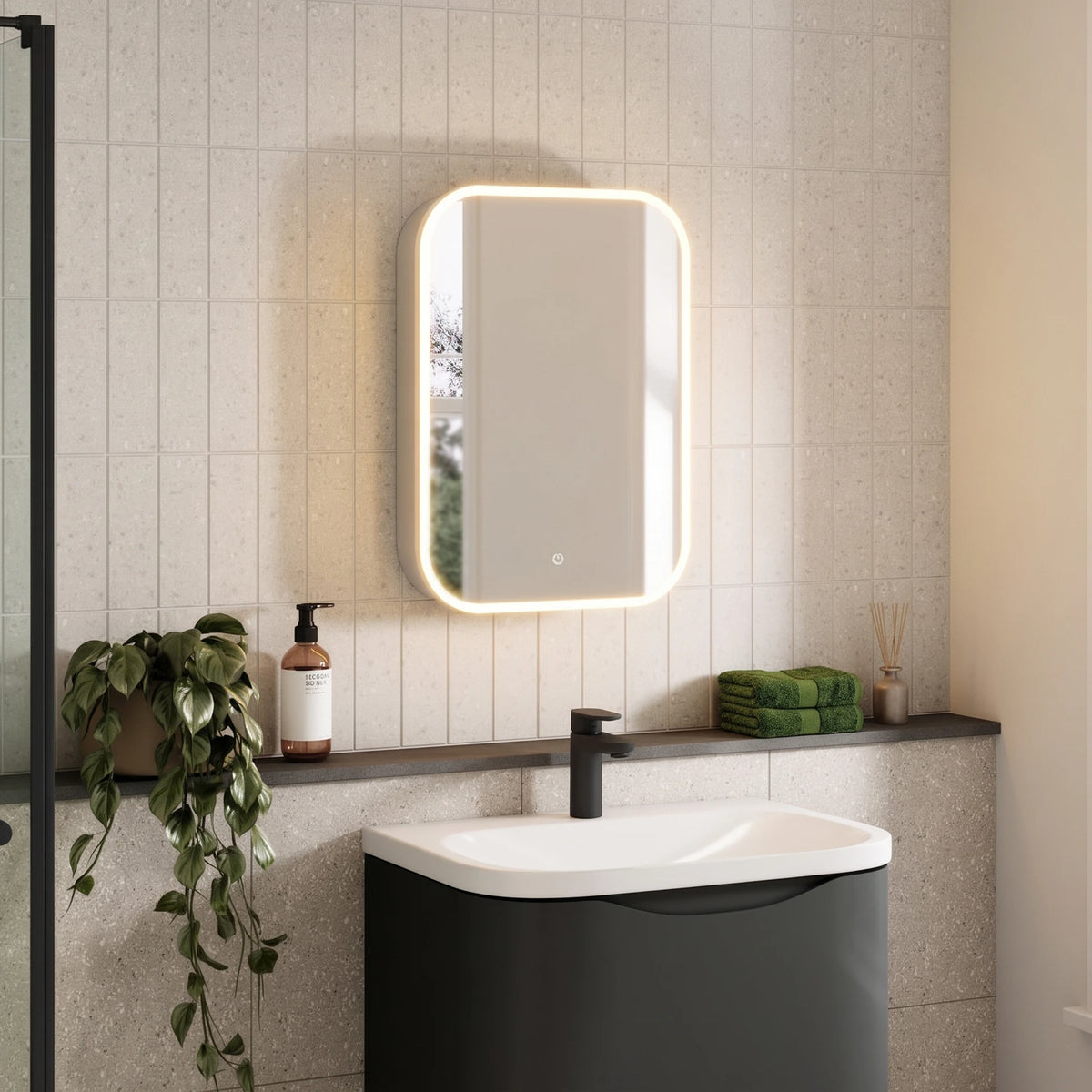 A modern bathroom scene featuring a rectangular LED-backlit mirror mounted on a light beige tiled wall above a dark gray and white sink vanity. A small circular touch control is visible at the bottom center of the mirror. #Size_500mm x 700mm