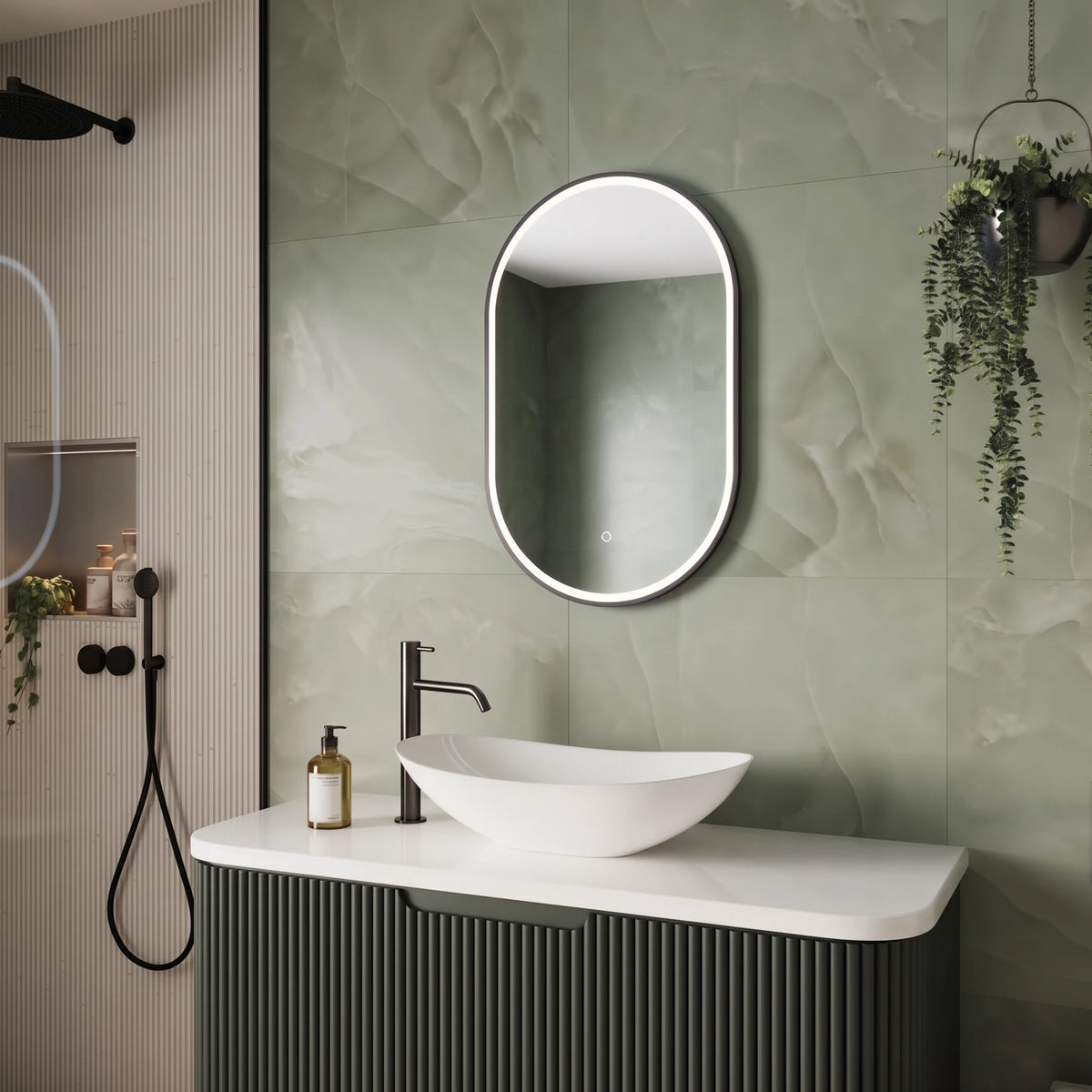 An oval LED-backlit bathroom mirror with a slim silver frame, mounted on a textured sage green tiled wall above a white vessel sink and a dark ribbed vanity cabinet. #Finish_Black