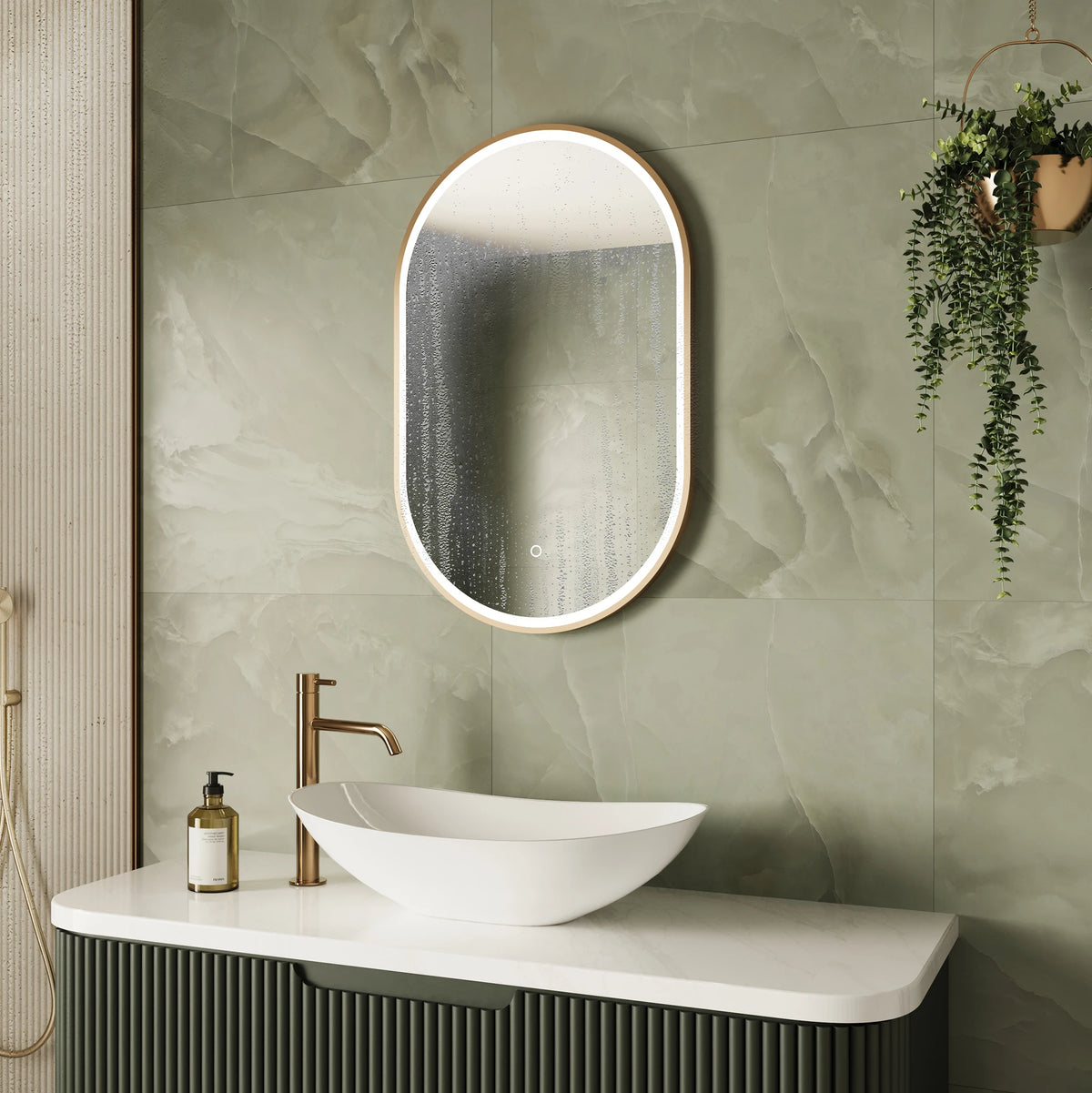 An oval LED-backlit mirror with a gold frame hangs on a green marble-look tiled bathroom wall above a white vessel sink and a modern dark green vanity cabinet. #Finish_Brushed Brass