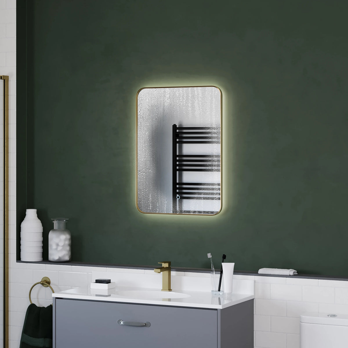 A rectangular wall mirror with a warm white LED light glow around its edge, mounted on a dark green bathroom wall above a white vanity. #Finish_Brushed Brass