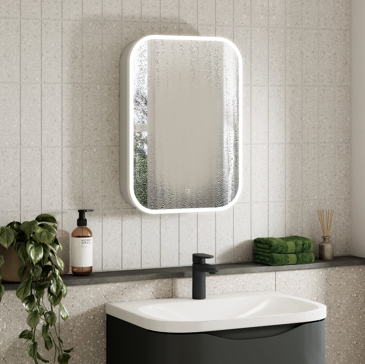 A modern bathroom scene featuring a rectangular wall-mounted mirror with a white frame and integrated LED lighting, mounted on a light beige tiled wall above a white and dark gray sink. #Size_500mm x 700mm