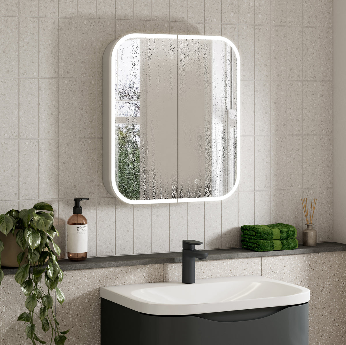 A modern bathroom scene featuring a rectangular wall-mounted mirror with a bright white LED light frame, set against light gray speckled tiles above a white and dark gray sink. A small plant, a bottle of hand wash, and folded green towels are arranged on the shelf below. #Size_600mm x 700mm