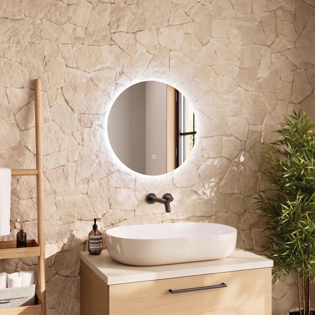 A round backlit LED vanity mirror mounted on a textured stone wall above a white vessel sink and light wood vanity cabinet, with a wooden towel ladder and green plant in a bathroom setting. #Size_⌀500mm