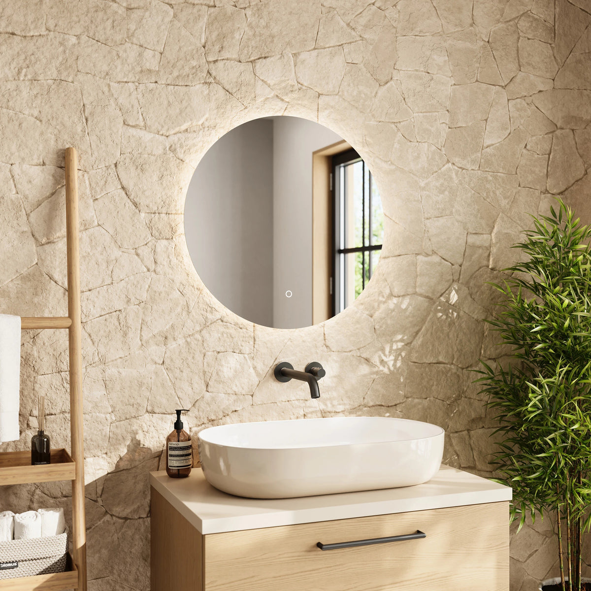 A round wall-mounted mirror with a black frame and integrated lighting, installed above a white vessel sink on a light wood vanity in a bathroom with a textured beige stone wall. #Size_⌀600mm