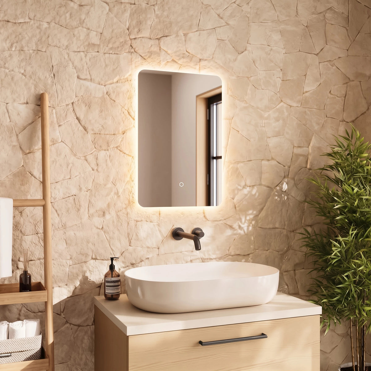 A modern bathroom scene featuring a rectangular LED-backlit mirror mounted on a textured beige stone wall above a light wood vanity with a white vessel sink. A wooden ladder shelf with toiletries stands on the left, and a green potted plant is on the right. #Size_400mm x 600mm