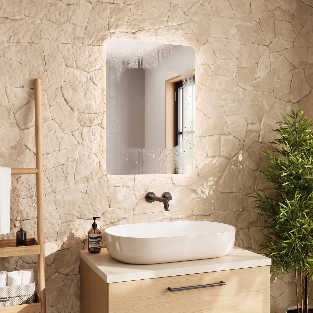 A modern bathroom scene featuring a rectangular LED-backlit mirror with a rounded top, mounted on a textured beige stone wall above a light wood vanity with a white vessel sink. #Size_500mm x 700mm