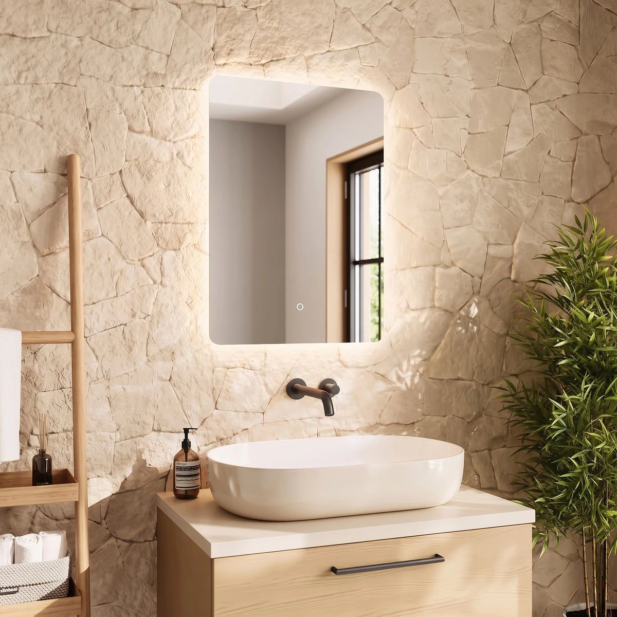 A modern bathroom vanity setup featuring a white oval vessel sink on a light wood cabinet, set against a textured beige stone wall with an illuminated rectangular mirror above. A wooden towel ladder, a dark bottle, and a green potted plant are visible in the styled room. #Size_500mm x 700mm