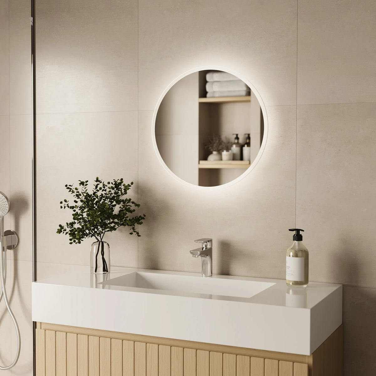 A modern bathroom vanity setup with a rectangular white sink, a light wood cabinet, a clear glass vase with greenery, and a pump bottle on the counter. Above the sink is a round backlit mirror mounted on a beige tiled wall.