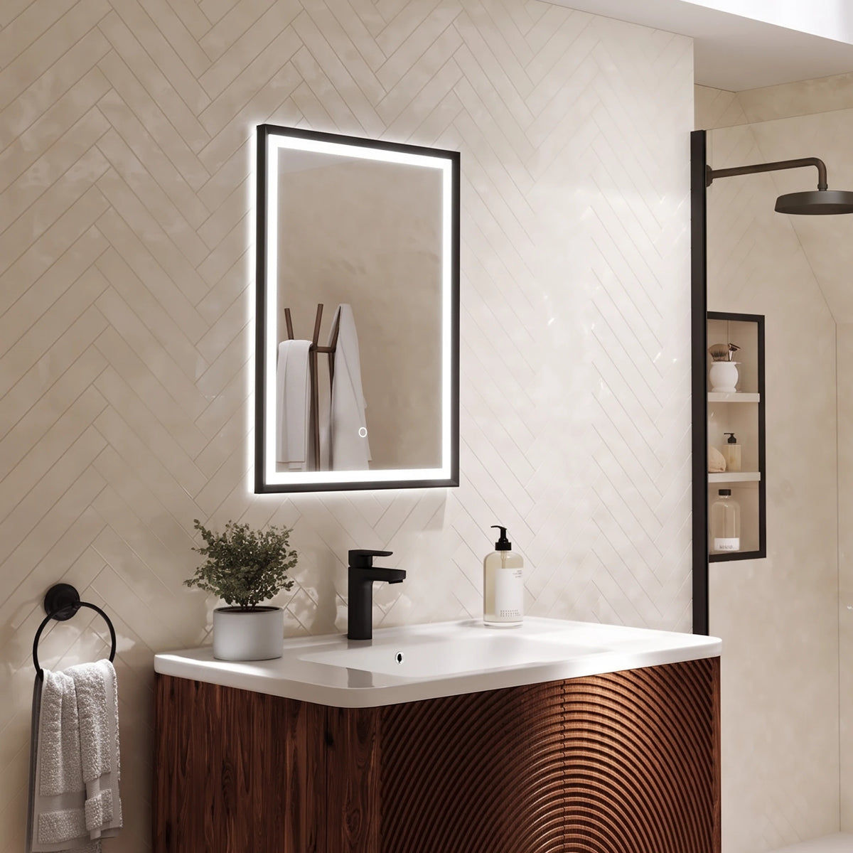 A modern bathroom vanity setup featuring a rectangular LED-backlit mirror with a black frame, mounted on a beige herringbone tile wall above a white countertop and a dark wood cabinet with a curved front. A matte black faucet, a small potted plant, and a white towel are visible. #Finish_Black