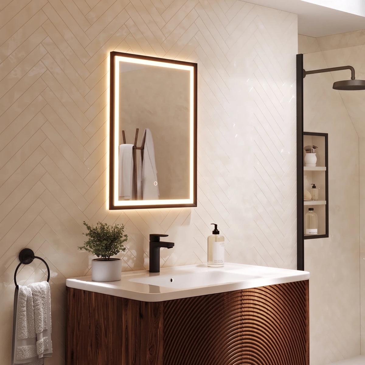 A modern bathroom vanity setup featuring a rectangular backlit LED mirror with a dark wood frame, mounted on beige herringbone tile. Below the mirror is a white countertop with a matte black faucet, a small potted green plant, and a white soap dispenser. #Finish_Black