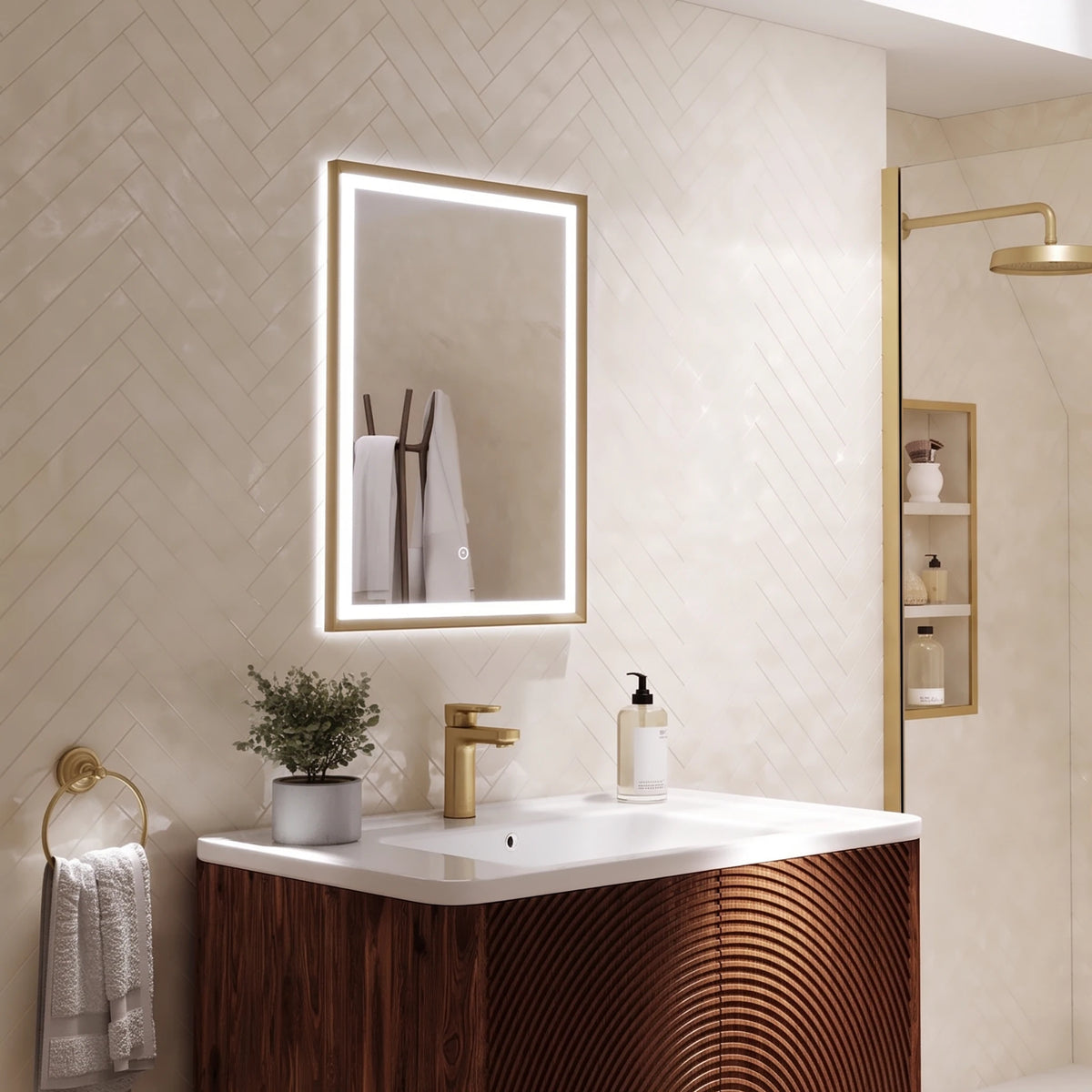 A modern bathroom vanity setup featuring a rectangular LED-backlit mirror with a gold frame, a white countertop, a gold faucet, and a dark wood cabinet with a curved slatted front. The background consists of beige herringbone tile walls, with a shower area visible on the right. #Finish_Brushed Brass