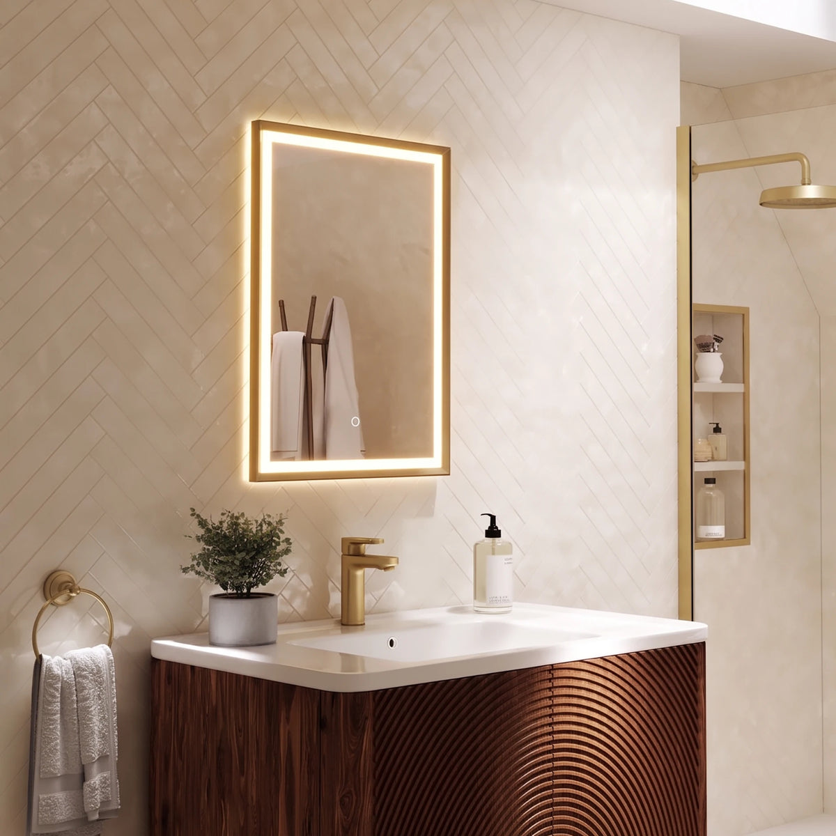 A modern bathroom vanity featuring a rectangular LED-backlit mirror with a gold frame, mounted on a beige herringbone tile wall above a white countertop and a dark wood cabinet. A gold faucet, white towels, and a small potted plant are visible in the scene. #Finish_Brushed Brass