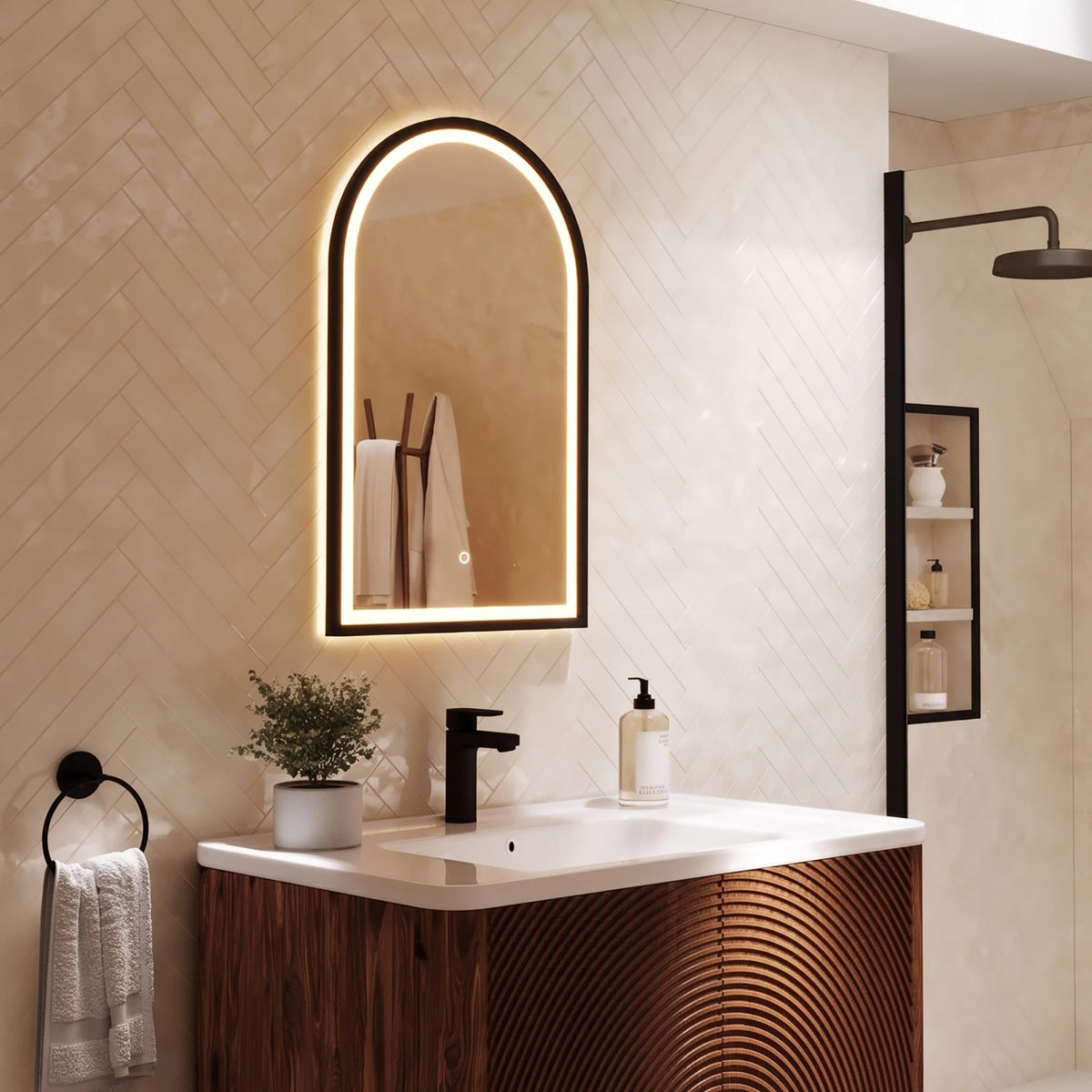 An illuminated arched wall mirror with a black frame hangs above a bathroom vanity. The scene includes a beige herringbone tile wall, a white countertop, a black faucet, a small potted plant, a towel ring with a white towel, and a shower fixture in the background. #Finish_Black