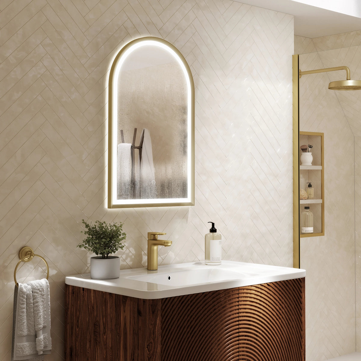 An illuminated arched bathroom mirror with a gold frame hangs above a white sink and a wood-look vanity in a modern beige bathroom. A gold faucet, towel ring, and shower area are visible, with a small plant and soap dispenser on the counter. #Finish_Brushed Brass