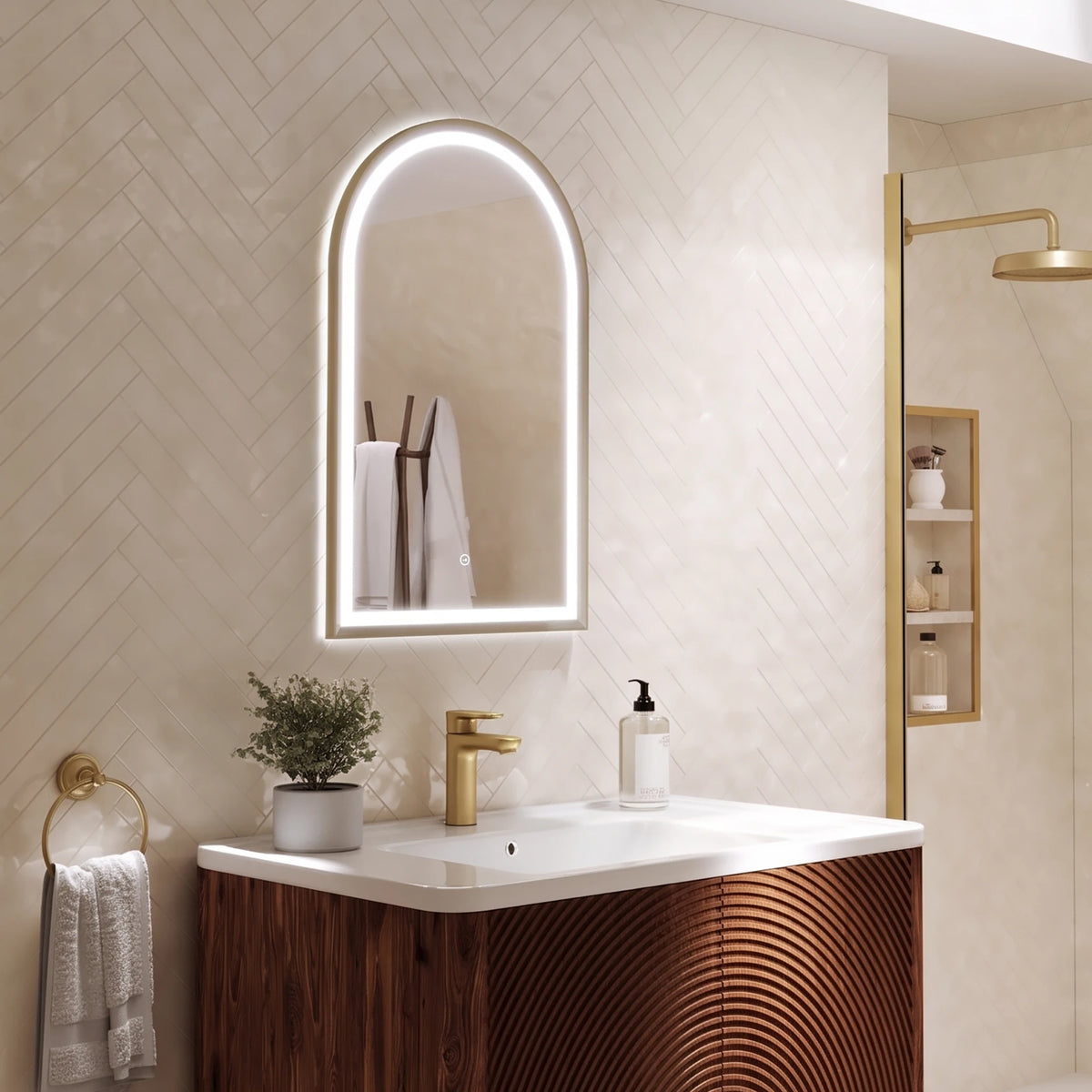 An illuminated arched LED bathroom mirror with a slim gold-toned frame, mounted on a beige herringbone tile wall above a modern vanity with a white countertop and wood-look front. #Finish_Brushed Brass