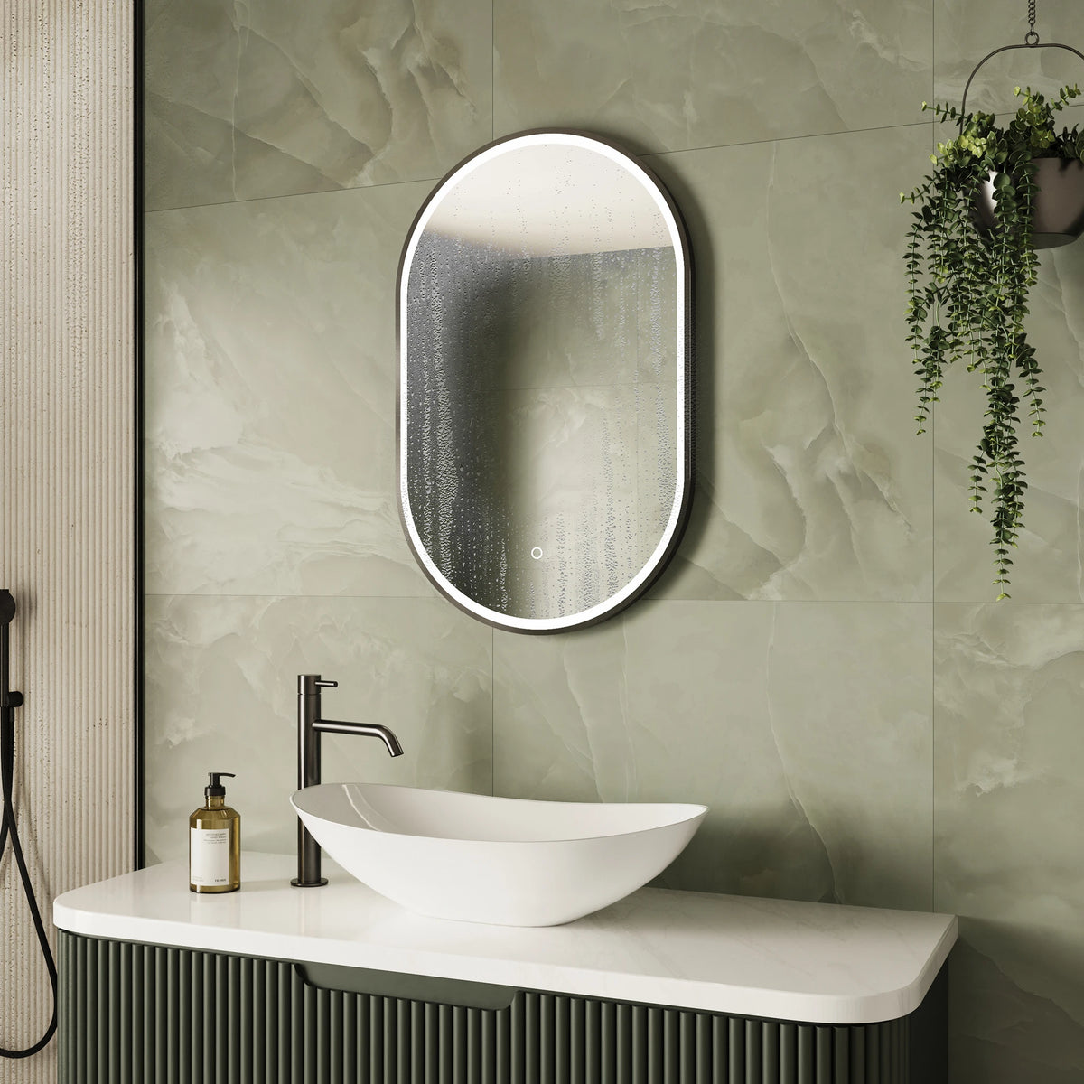 An oval LED-backlit bathroom mirror with a slim black frame, mounted on a light green marble-look tiled wall above a white vessel sink and a dark green ribbed vanity cabinet. #Finish_Black