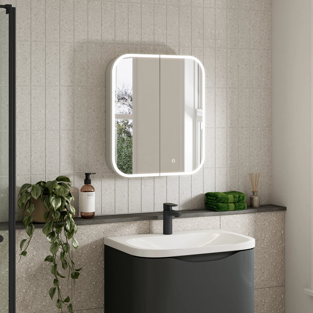 A modern bathroom scene featuring a rectangular wall-mounted mirror with a glowing white LED light border, installed above a white and dark gray sink. The background consists of light beige textured wall tiles, with a dark shelf holding a green plant, a brown bottle, and a folded green towel. #Size_600mm x 700mm