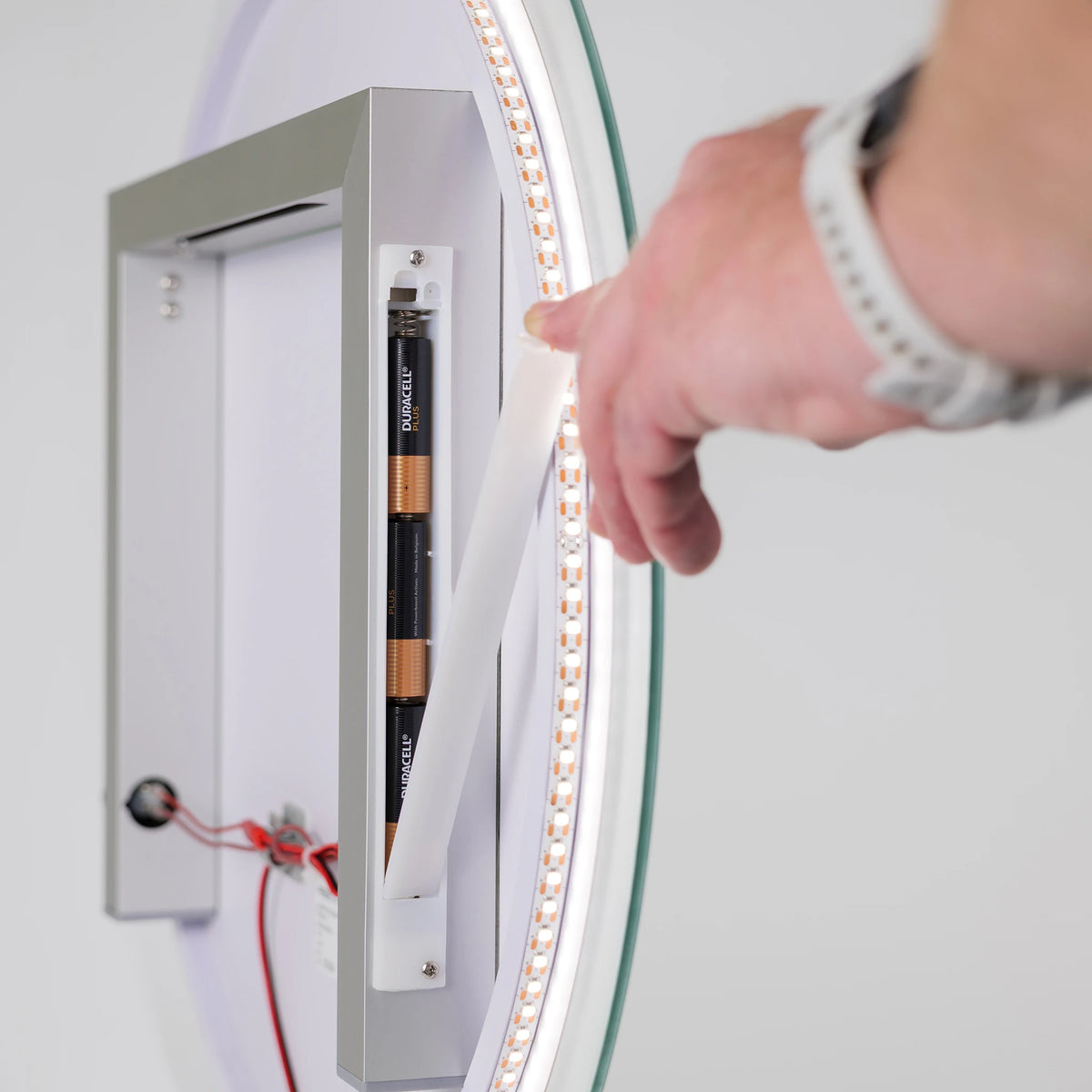 A close-up of a hand installing or adjusting a white flexible light strip around the inner edge of a circular glass mirror, with exposed battery compartments and wiring visible inside a silver frame.