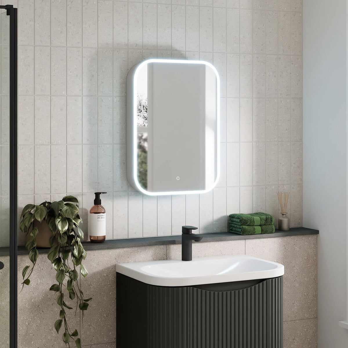 A modern bathroom scene featuring a rectangular LED-backlit mirror mounted on a light beige tiled wall above a dark gray ribbed vanity with a white sink. A black faucet, a potted trailing green plant, and a folded green towel are also visible. #Size_500mm x 700mm